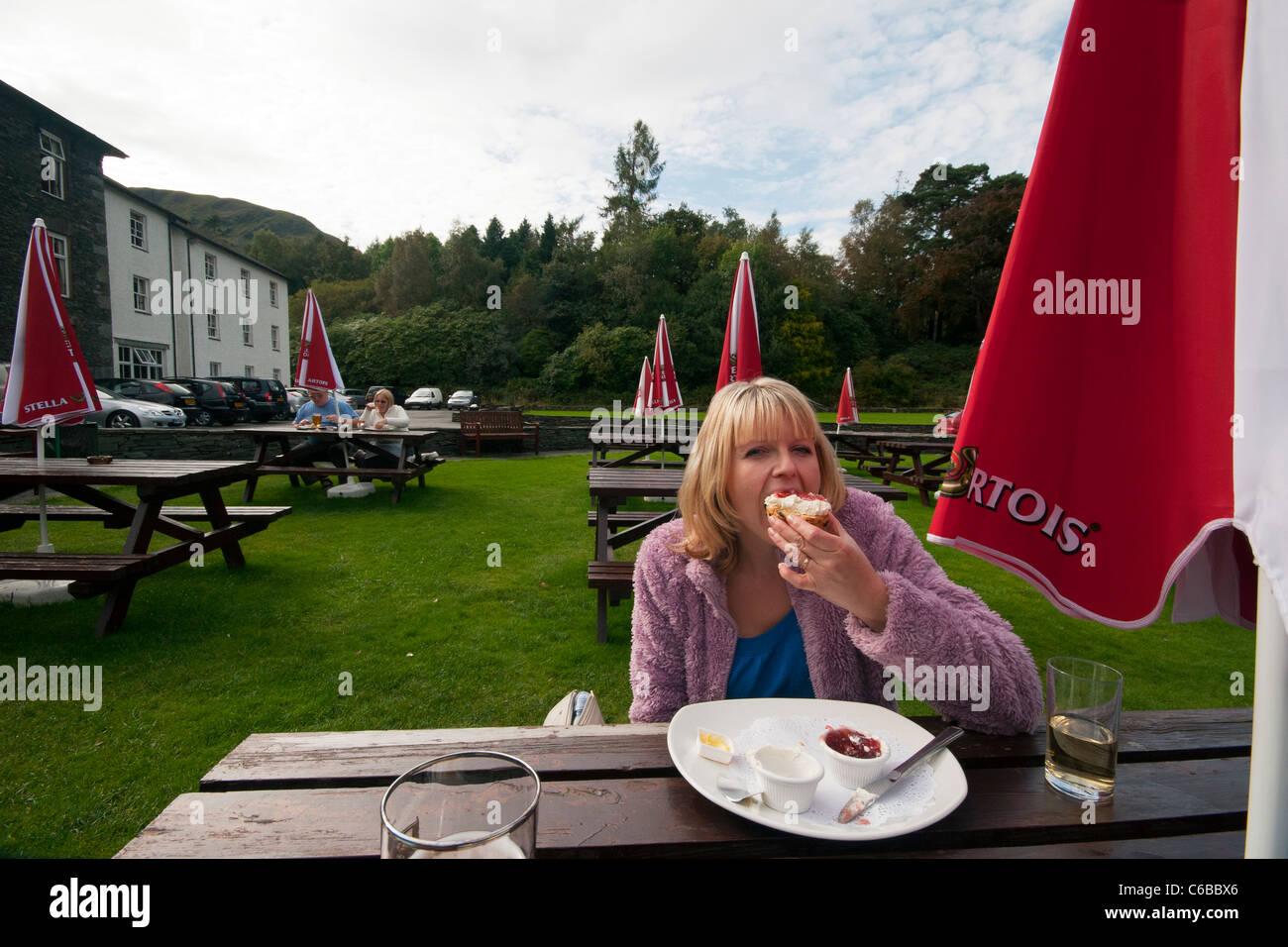 Lady stuffing face with cream tea scone with jam eating last bit on pub bench with parasol pub garden grounds Stock Photo