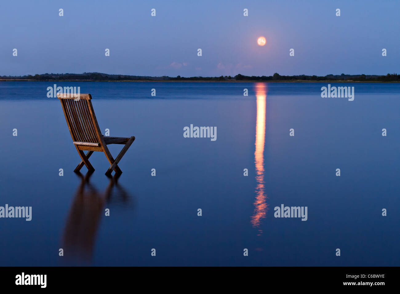 Singular chair in calm water facing the land in the horizon. With rising orange moon reflected in the blue water Stock Photo