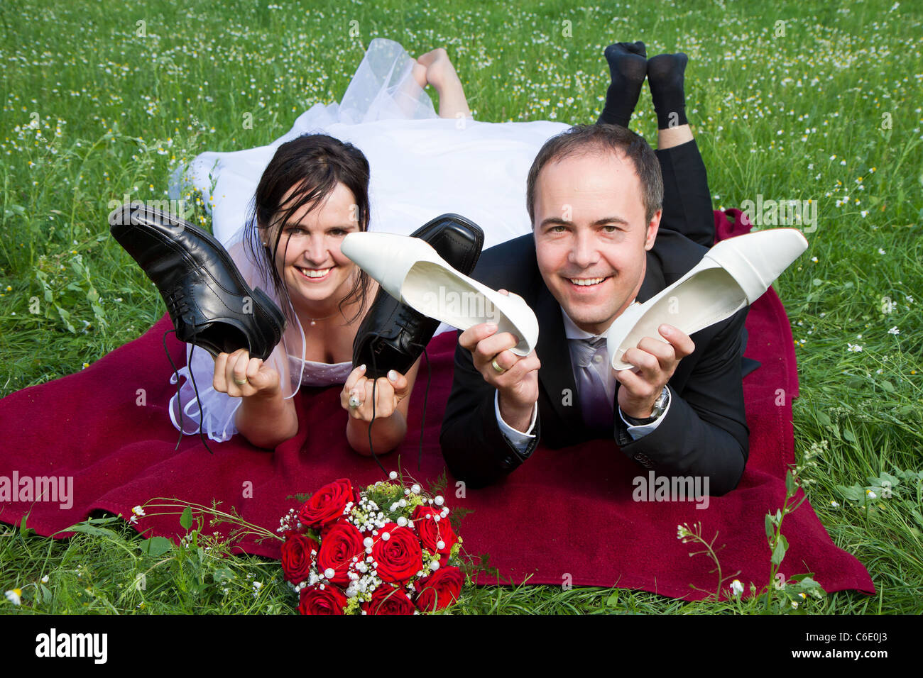 newly wed couple with wedding gown dark suit and red bridal bouquet the groom and bride lie on a meadow having fun Stock Photo