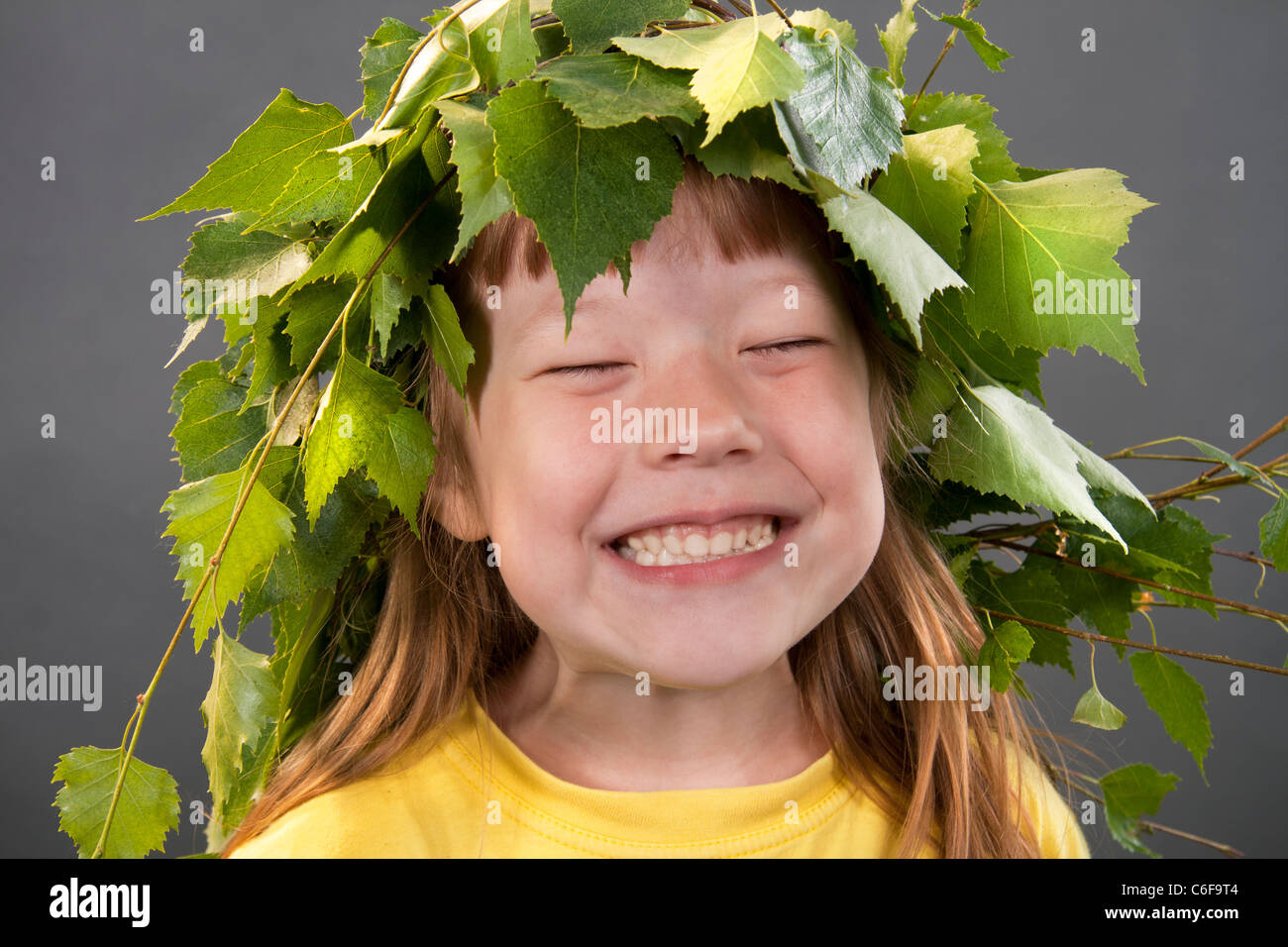 Cheerful little girl with a wreath on a head from birch leaves on a gray background Stock Photo