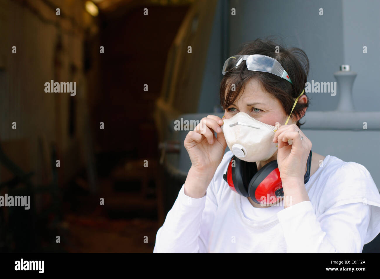 A young woman putting on a face mask, ear defenders and safety goggles for protection whilst working. Stock Photo