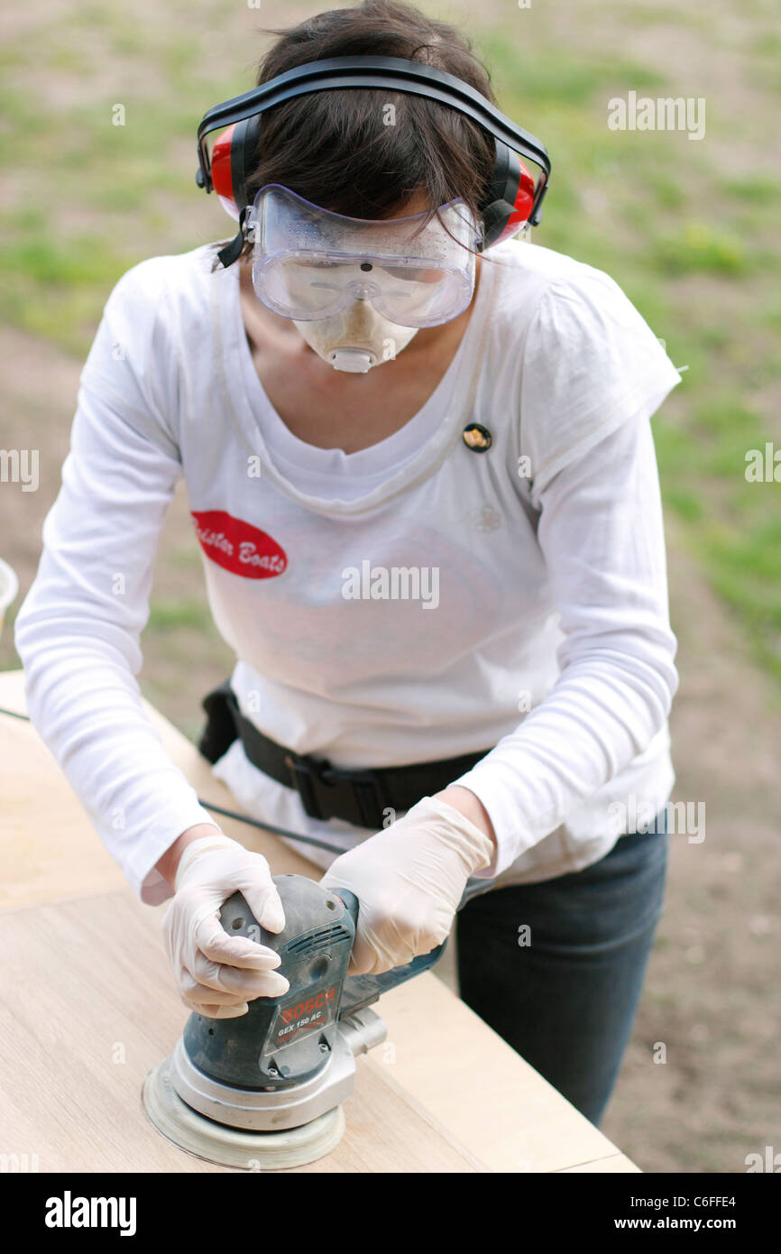 A woman sanding a wooden door at trestles, wearing ear defenders, safety goggles, latex gloves and a face mask for protection. Stock Photo