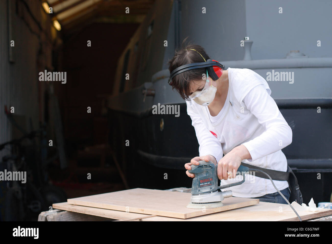 A woman sanding a wooden door at trestles, wearing ear defenders, safety goggles, latex gloves and a face mask for protection. Stock Photo