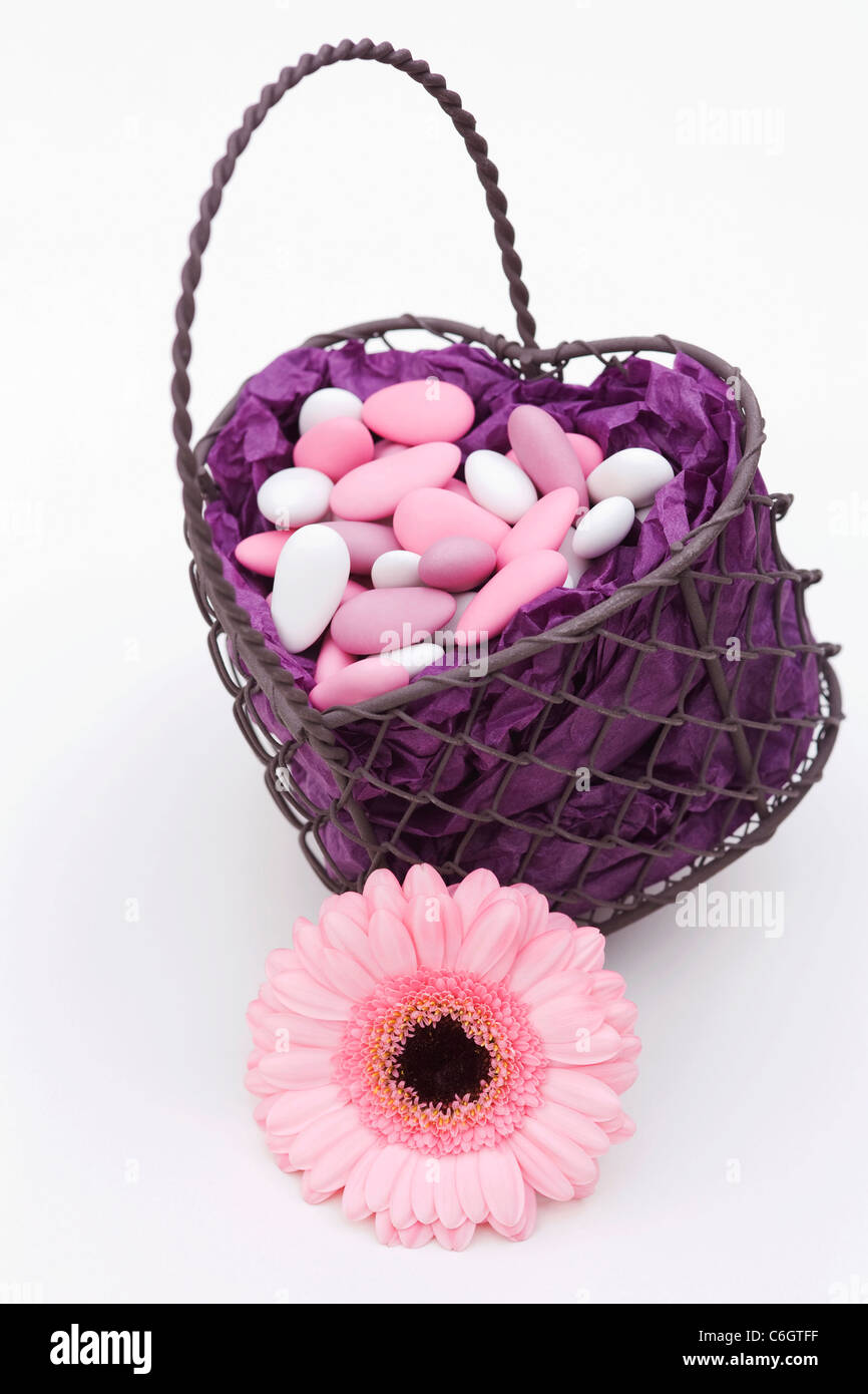 Sugared almonds in a heart shaped basket with a pink Gerbera on a white background. Stock Photo