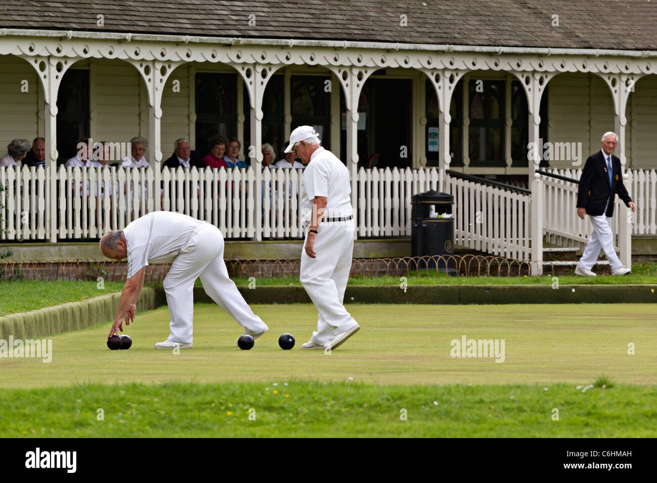 Senior gentlemen playing flat lawn green bowls picking up bowls in UK while other bowling club members look on appreciatively Stock Photo