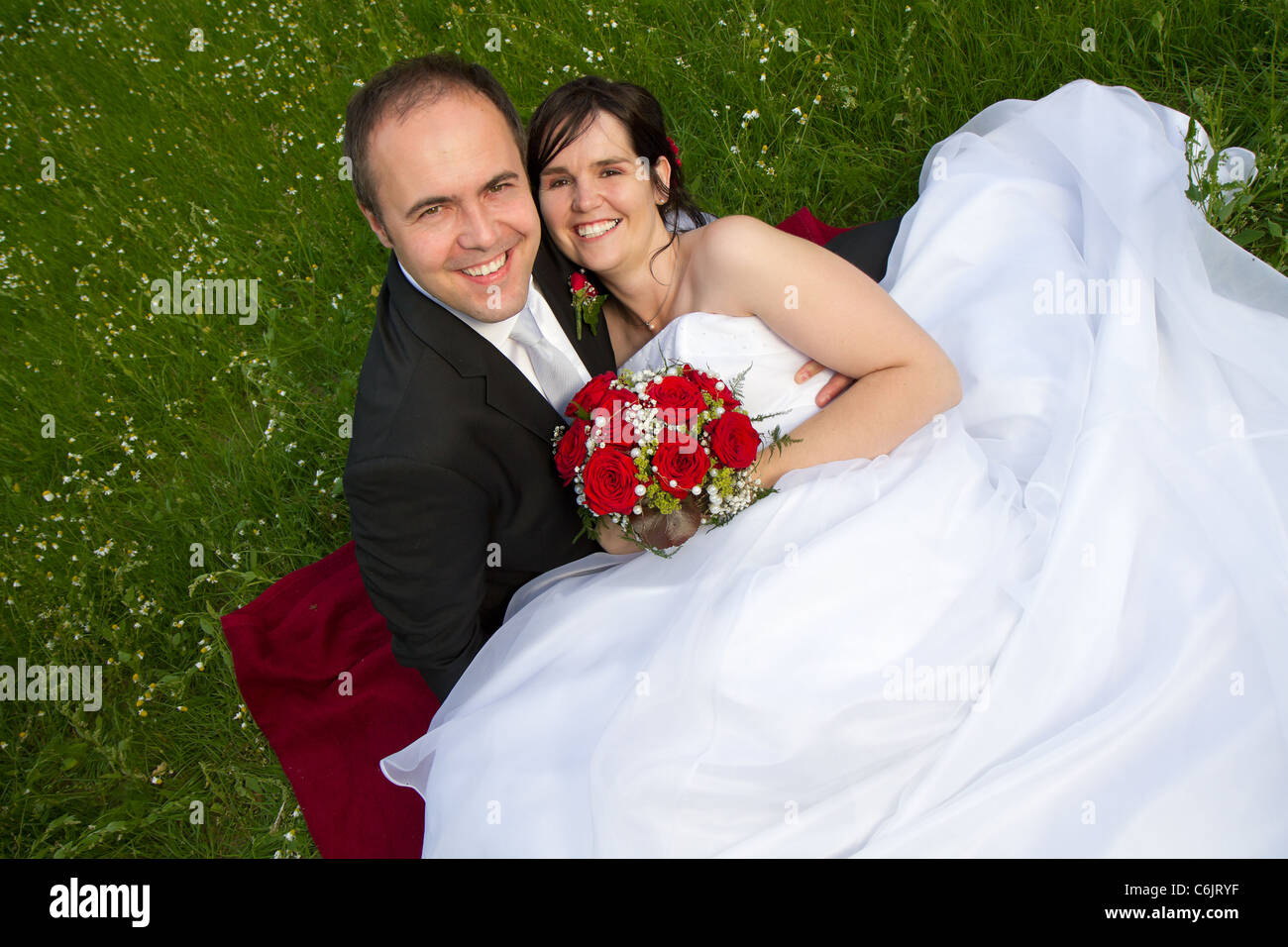 classical newly wed couple with wedding gown dark suit and red bridal bouquet the groom and bride lie on a meadow Stock Photo