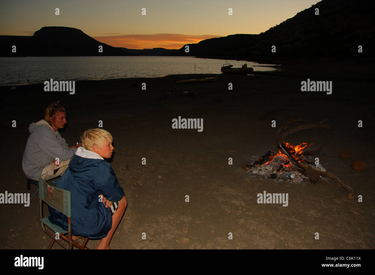 Campers sitting around a fire at the  edge of a dam at sunset Stock Photo