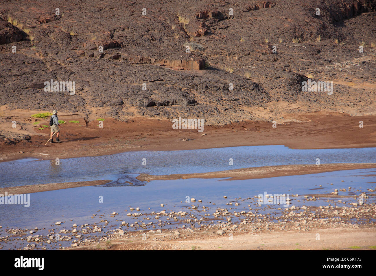 Lone hiker walking with walking stick along a shallow river Stock Photo