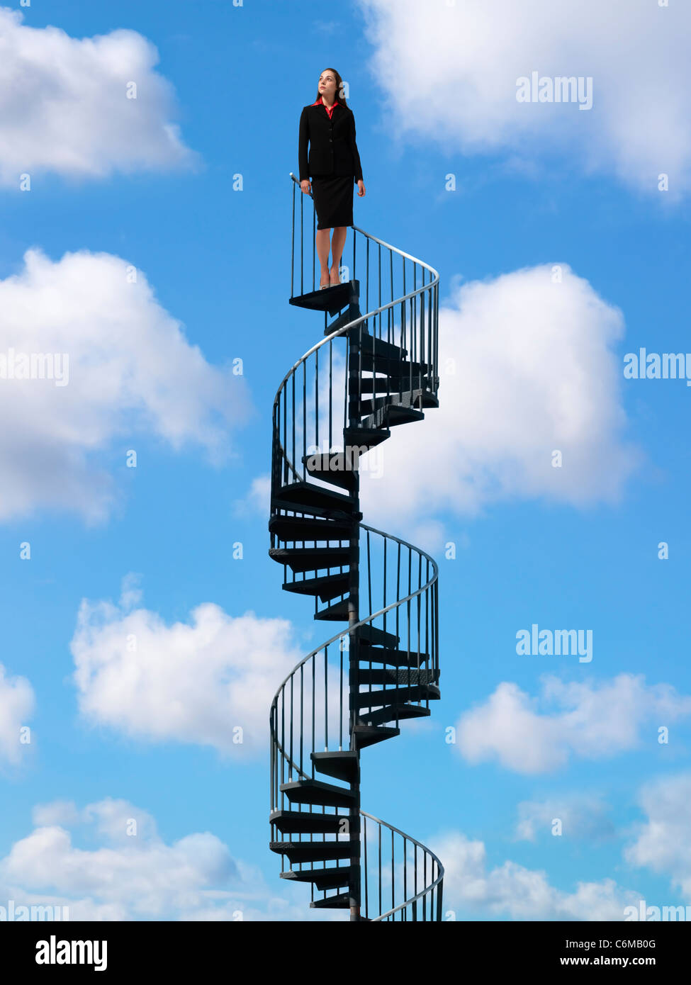 Woman standing at the top of metal spiral staircase rising through bright blue sky with puffy white clouds Stock Photo