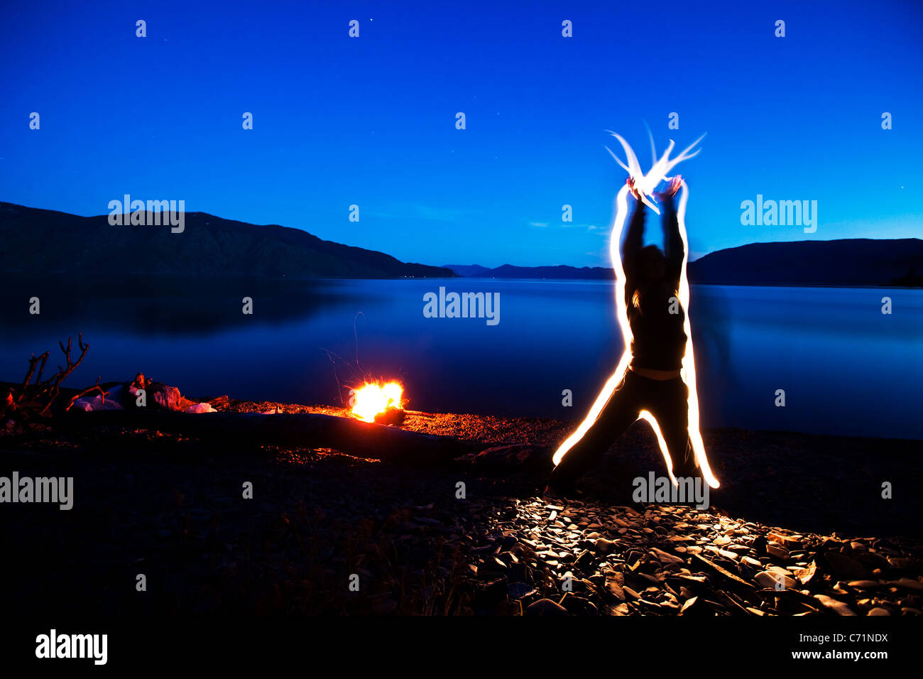 A woman sends energy through her body and out her hands at sunset next to a campfire and lake in Idaho. This light painting imag Stock Photo