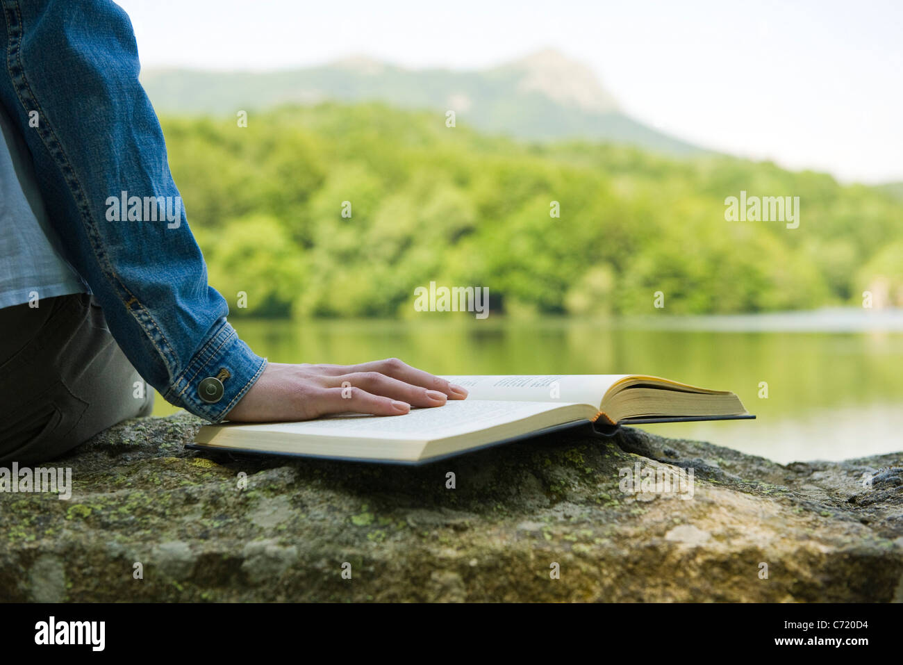 Woman sitting on rock by lake with book, cropped Stock Photo