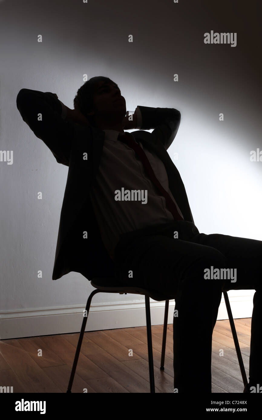 Silhouette of a young male sitting in a chair with his hands behind his head. Stock Photo