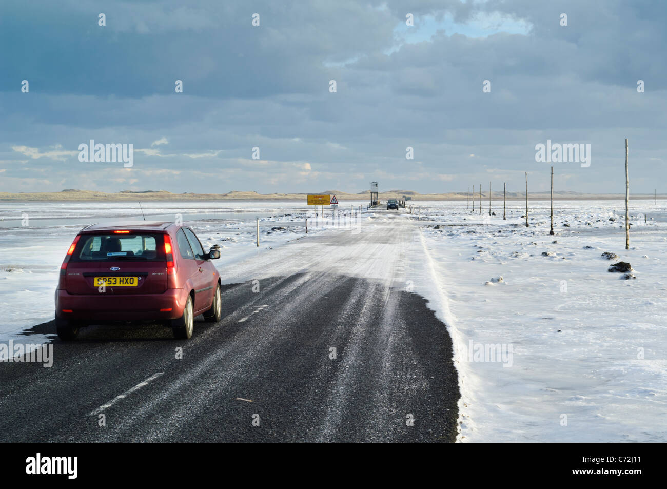 The tidal causeway road leading to Lindisfarne (Holy) Island, with the mudflats covered by ice. Car vehicle driving. Stock Photo
