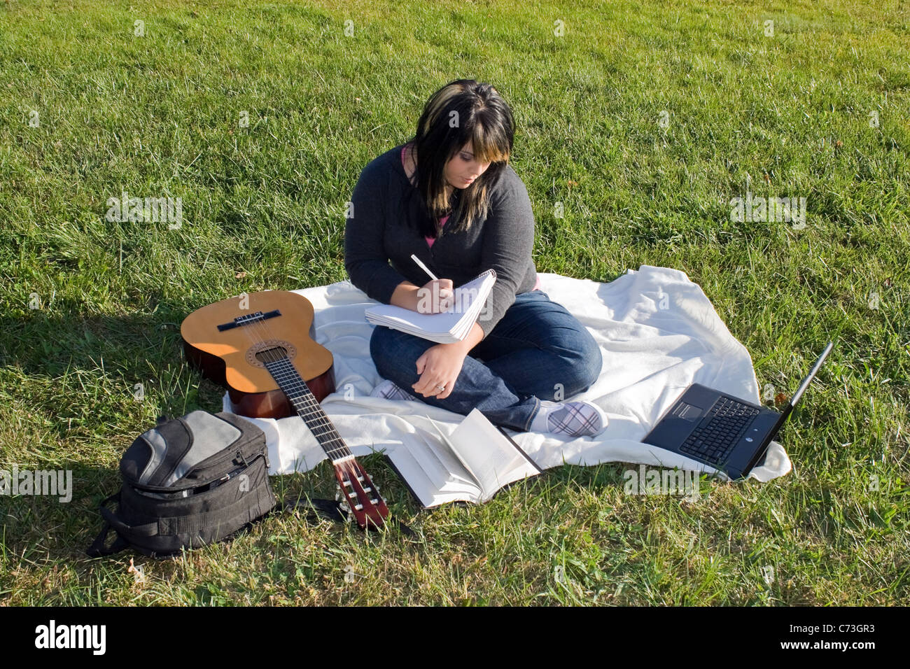 A young female singer or song writer with her guitar and computer outdoors in the grass. Stock Photo