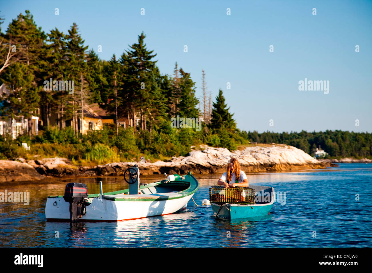 A lone woman sits in her boat in a small harbor at dusk after collecting lobsters from underwater traps. Stock Photo