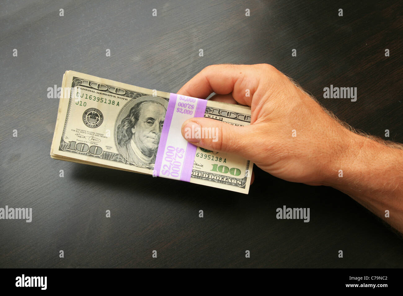 a mans hand holds two thousand dollars in hundred dollar bills on a dark wood table top Stock Photo