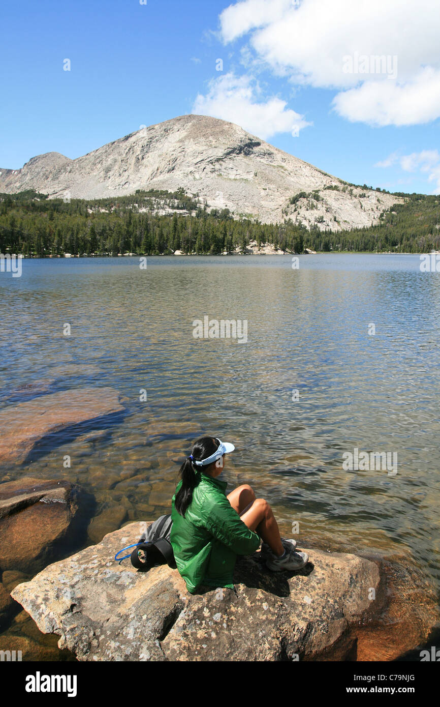 a woman in a green jacket sits on a rock by alpine Silas Lake in the Wind River Range, Wyoming Stock Photo