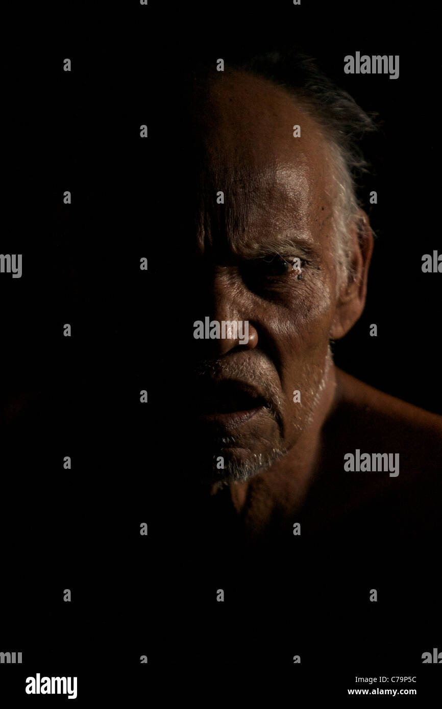 An elderly Indonesian man speaks animatedly form the shadows of his traditional hut. Stock Photo
