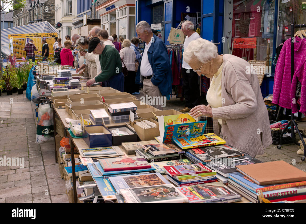 Mature female checks used books on an outdoor market stall, in the background bargain hunters check VHS tapes and magazines. Stock Photo