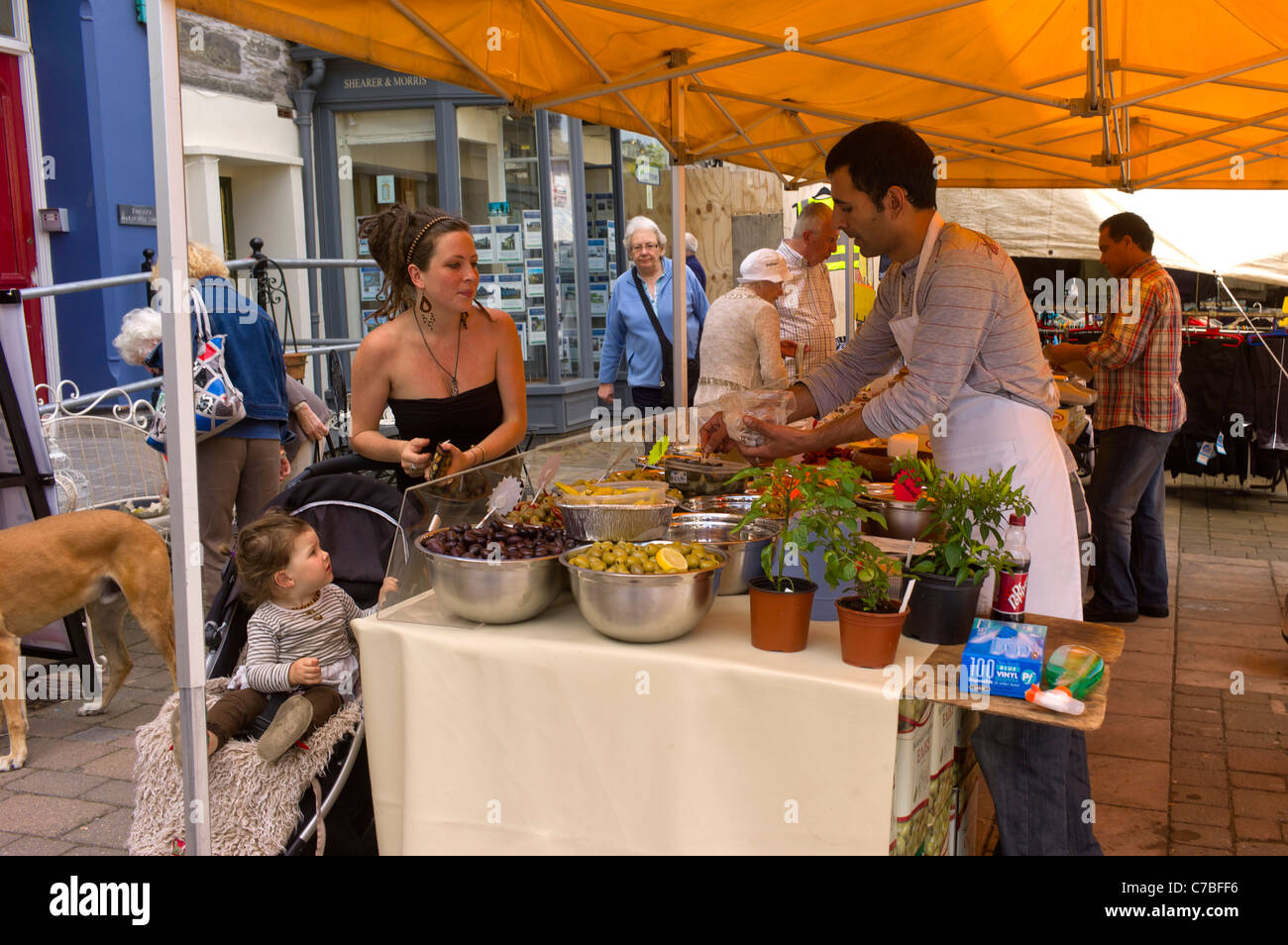 Attractive summery dressed mother with child in buggy market stall shops for salads and spices in town high street, busy scene. Stock Photo