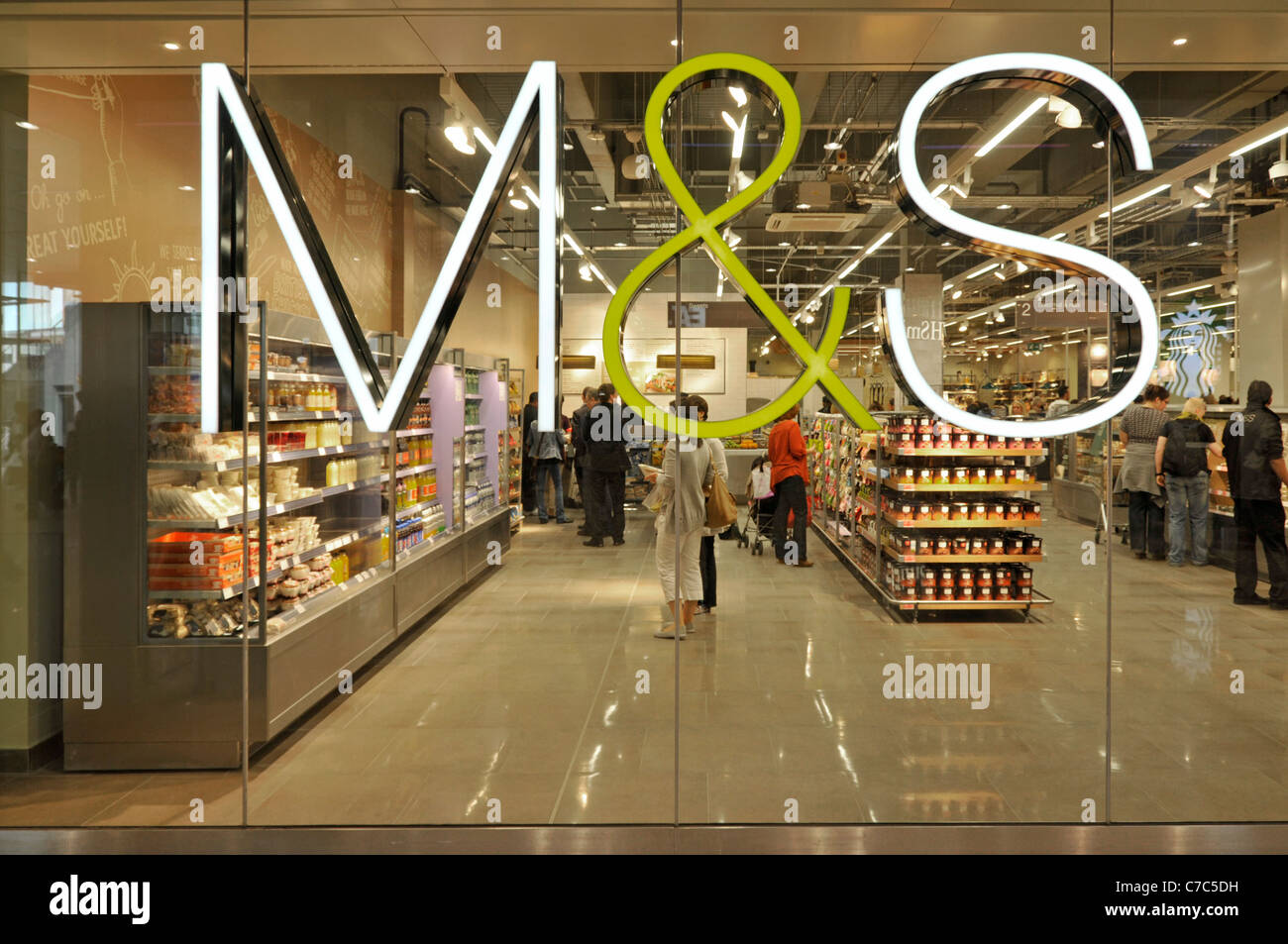 M&S retail business sign mounted on glass window of foodhall in Marks and Spencer store in Westfield Shopping Centre shoppers in food shop London UK Stock Photo
