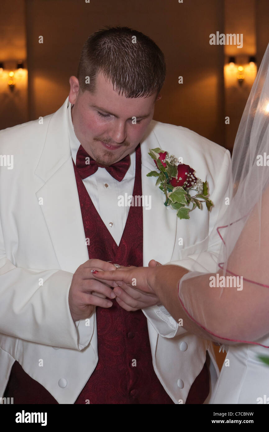 Groom dressed in white tuxedo putting wedding band on bride's finger during wedding ceremony Stock Photo