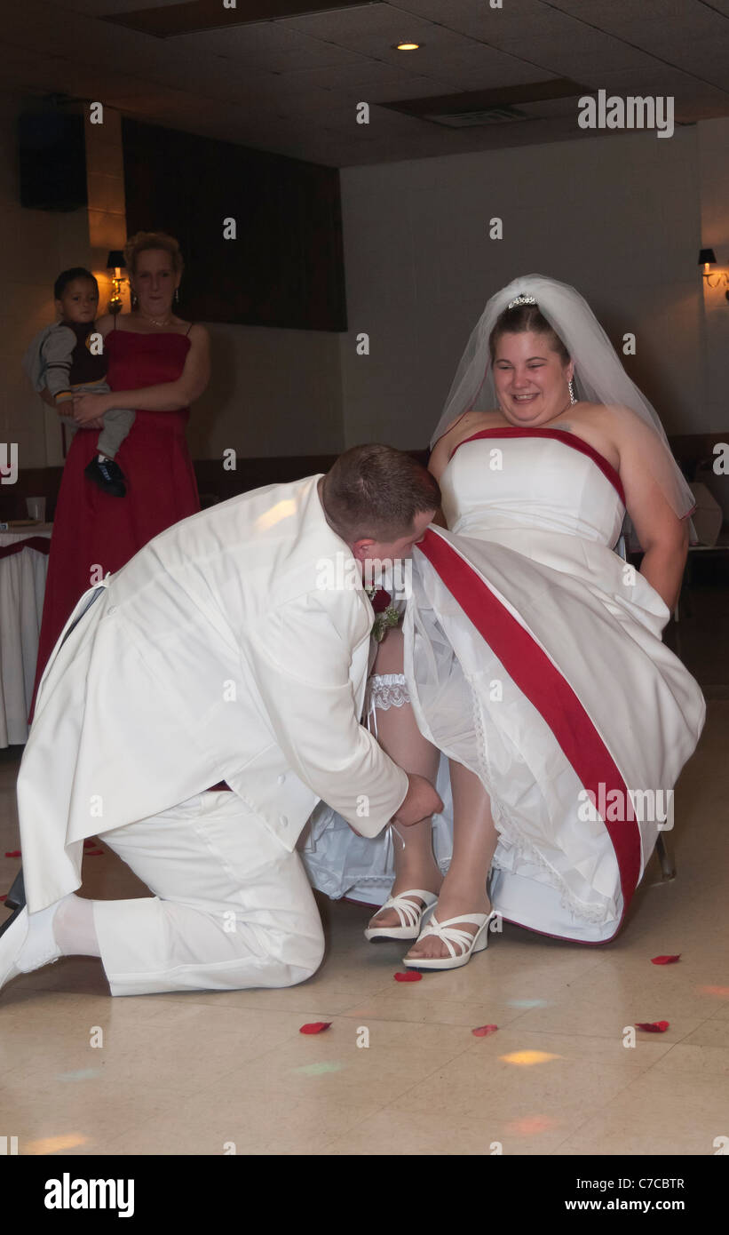Groom wearing white tuxedo taking off garter from bride's leg during wedding reception Stock Photo