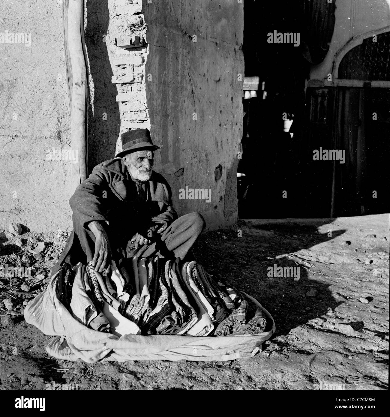 1950s. Historical picture showing bearded old man with hat sitting beside old building, selling blankets, Isfahan, Iran. Stock Photo