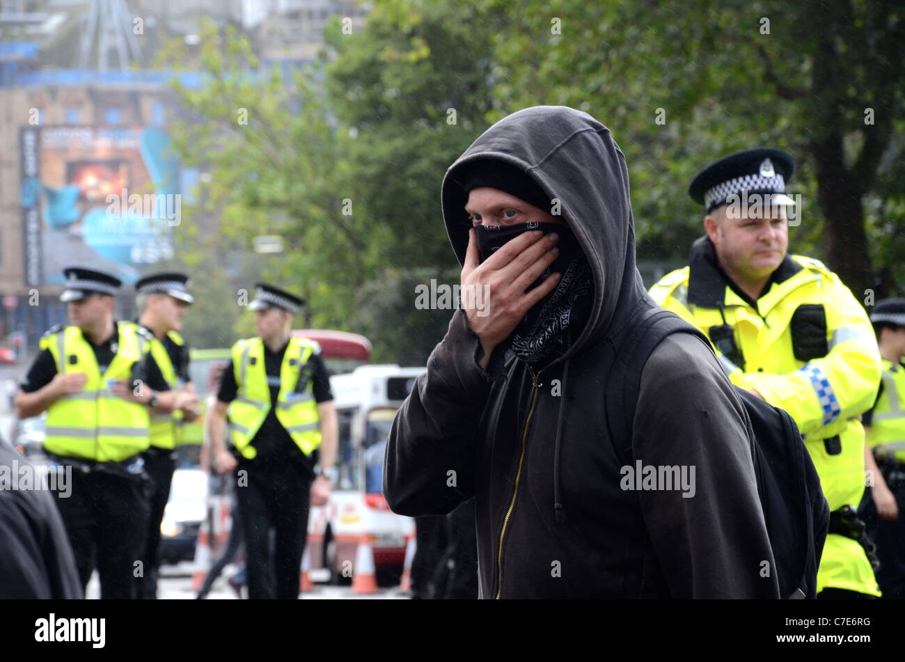 Hooded and masked protester walks along Princes Street in Edinburgh while the police look on in the background. Stock Photo