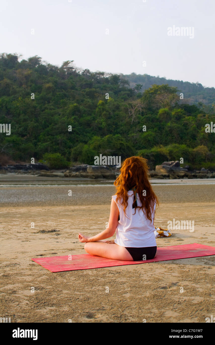 A young woman doing a yoga meditation pose on a beach in morning light ...