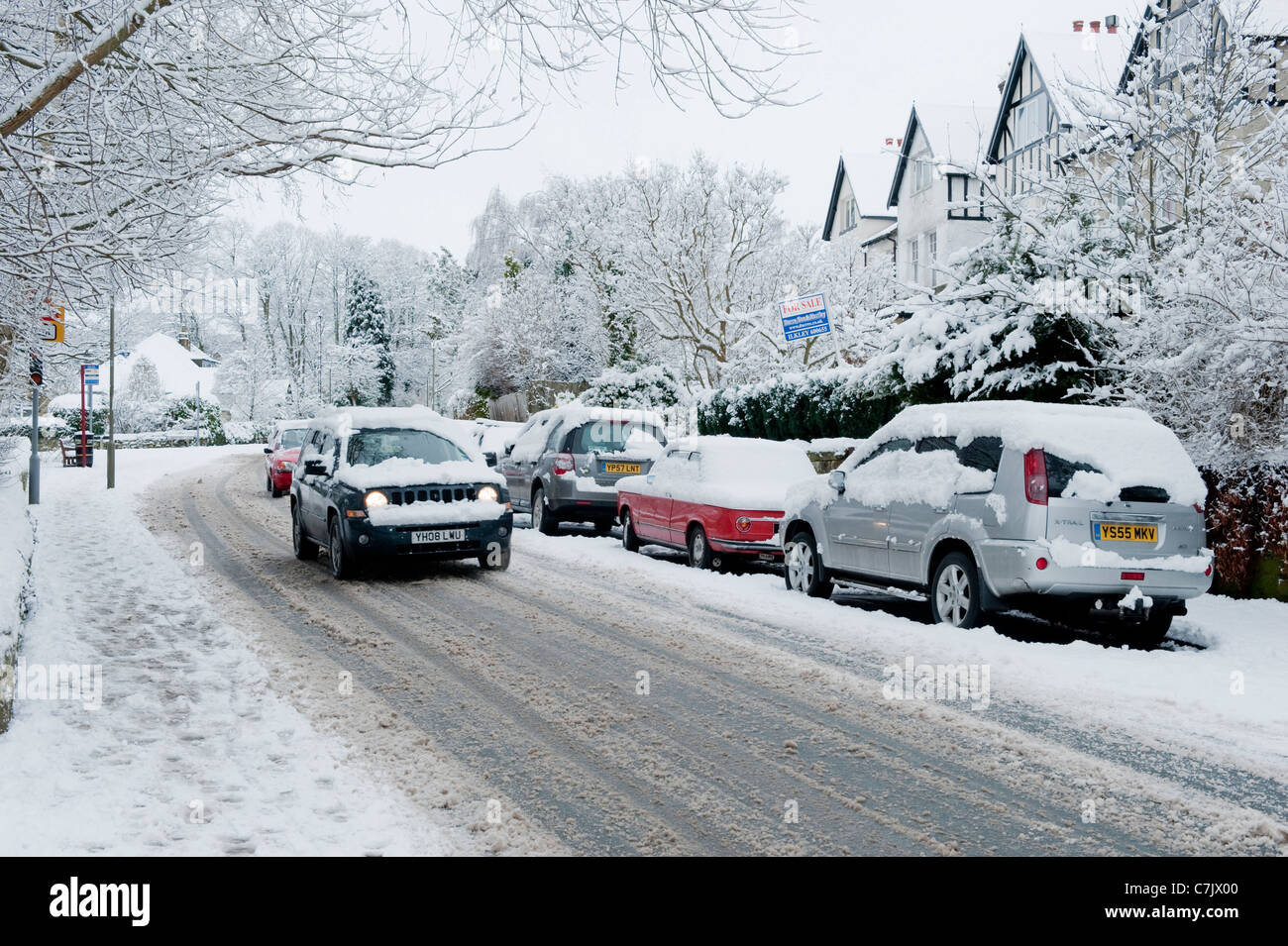 Winter village street (snowy driving conditions, Jeep traveling, cars parked on road, snow-covered pavement) - Burley in Wharfedale, England, GB, UK. Stock Photo