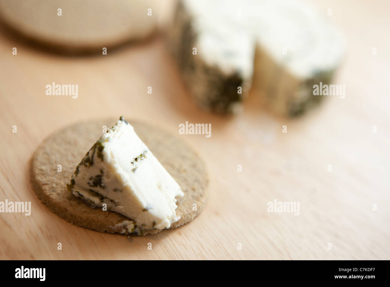 An oatcake with a slice of garlic and herb cream cheese, with oatcakes and cheese in the background against a wooden surface Stock Photo