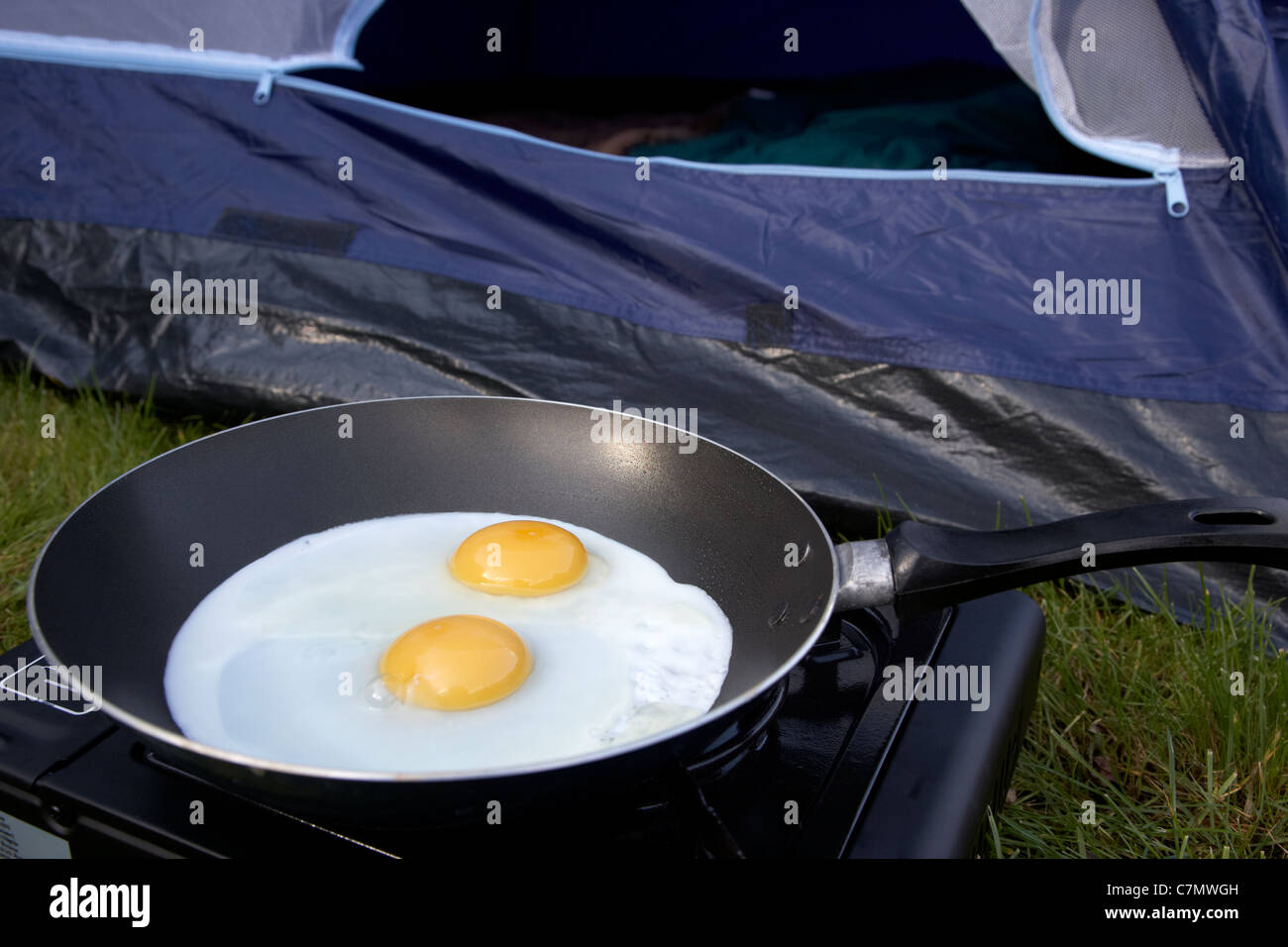 cooking two fried eggs on a small portable gas camping stove in front of the open door of a dome tent Stock Photo