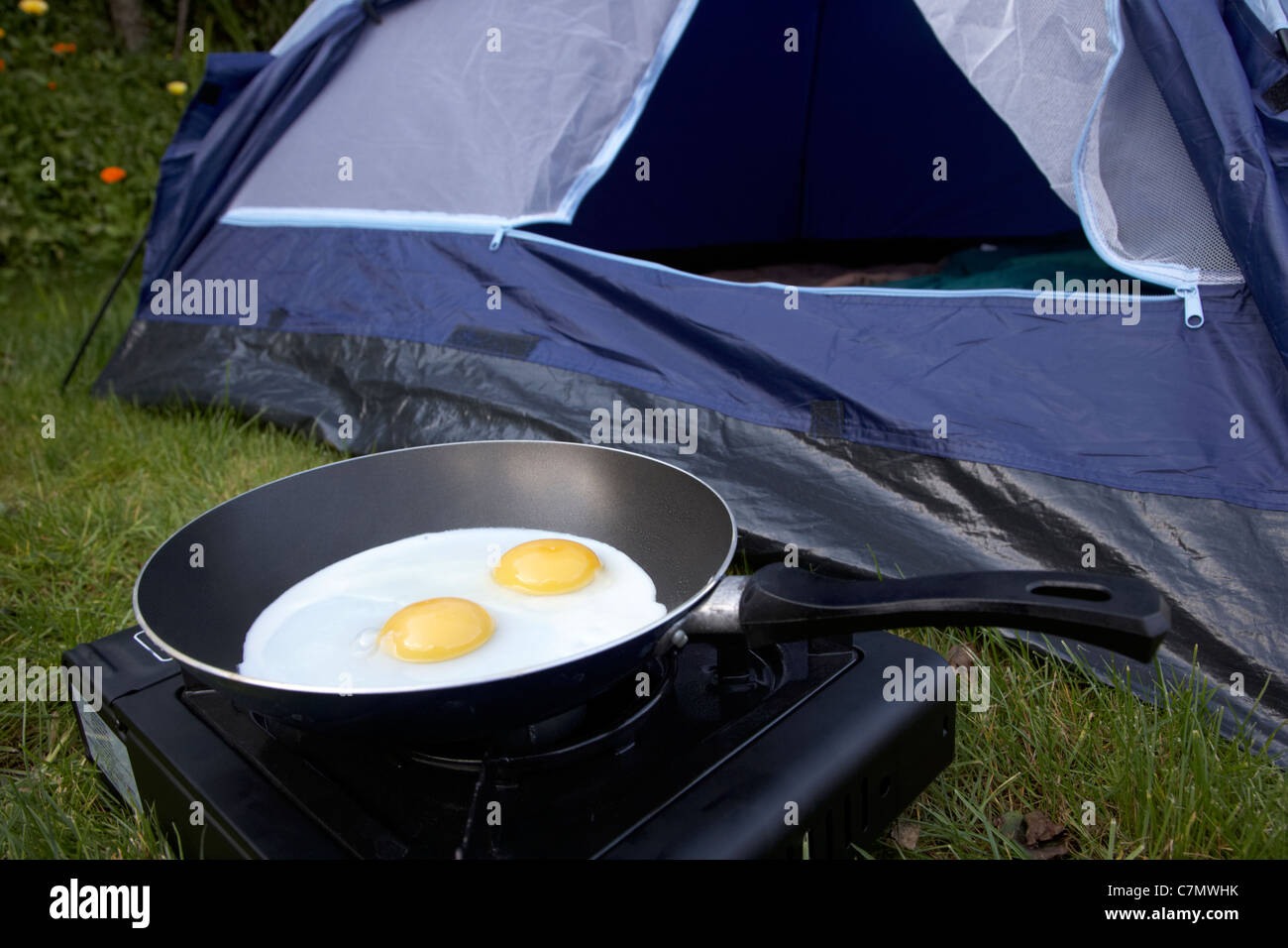 cooking two fried eggs on a small portable gas camping stove in front of the open door of a dome tent Stock Photo
