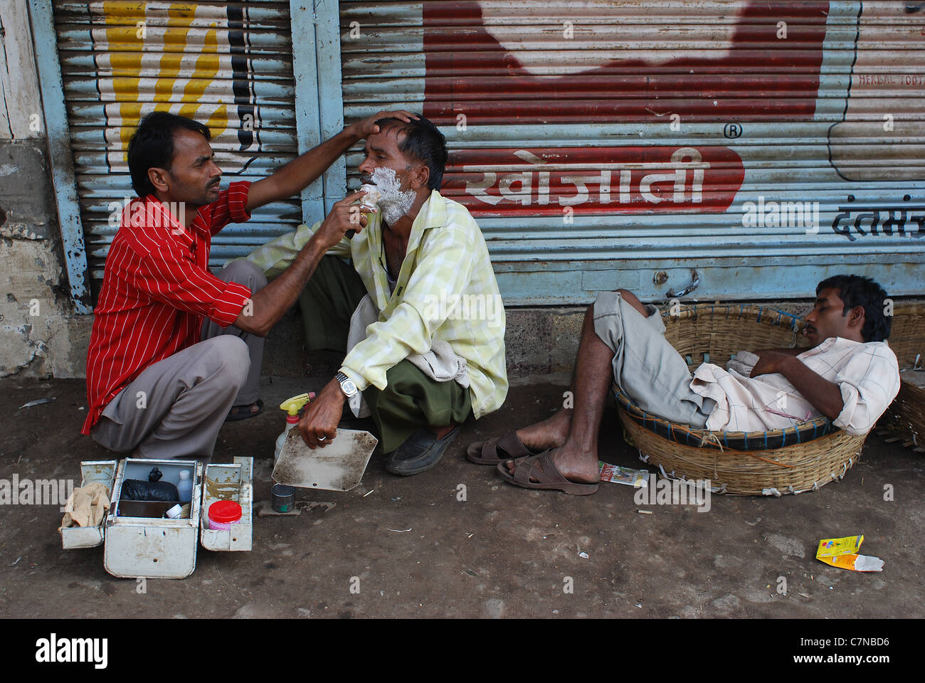 A barber is shaving a man in the street while another one is sleeping ( India) Stock Photo