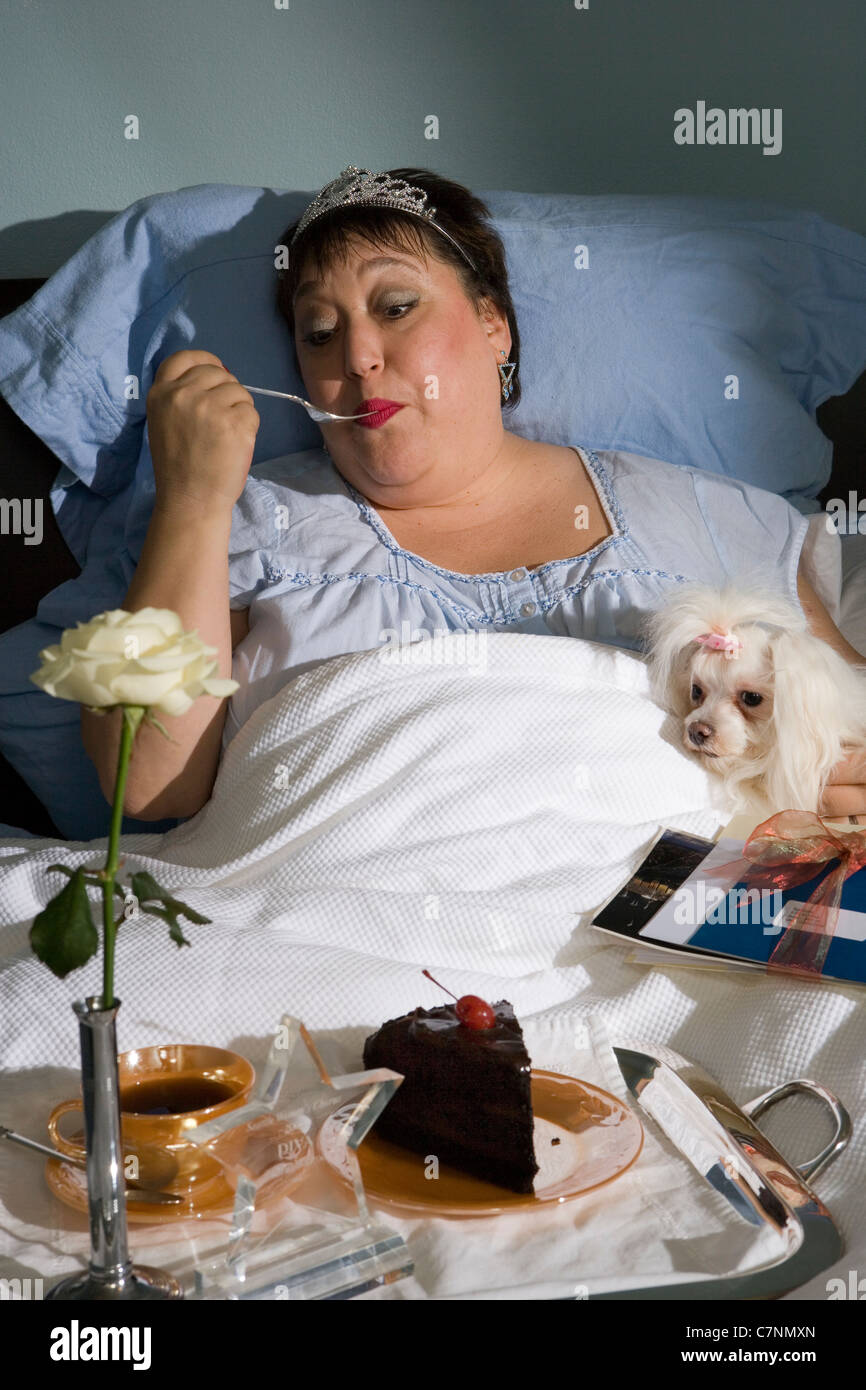 Diva's Breakfast in Bed. A larger than life woman dining on chocolate cake in bead with her beloved poodle looking on. Stock Photo