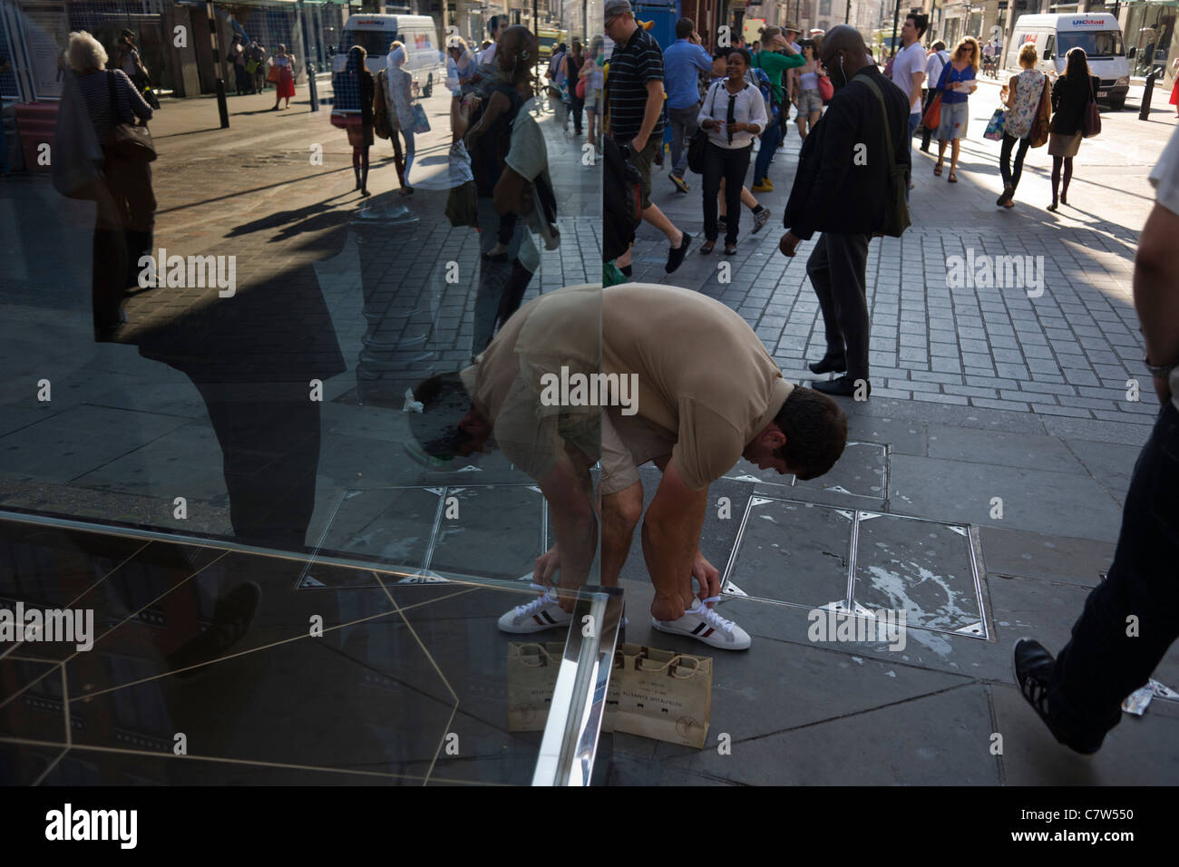 Young man reflected in glass who bends down to tie his trainer lace on a busy street corner near Covent Garden. Stock Photo