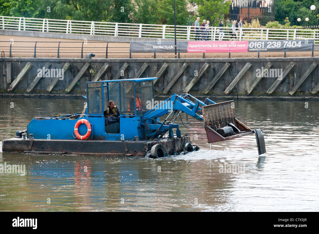Bucket loader on a barge, clearing floating debris on the Manchester Ship Canal, Salford Quays, Manchester, England, UK Stock Photo