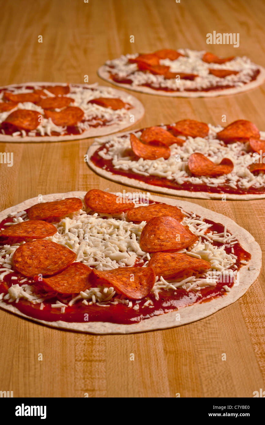 Four uncooked pepperoni and cheese tortilla pizzas on a long wooden surface Stock Photo