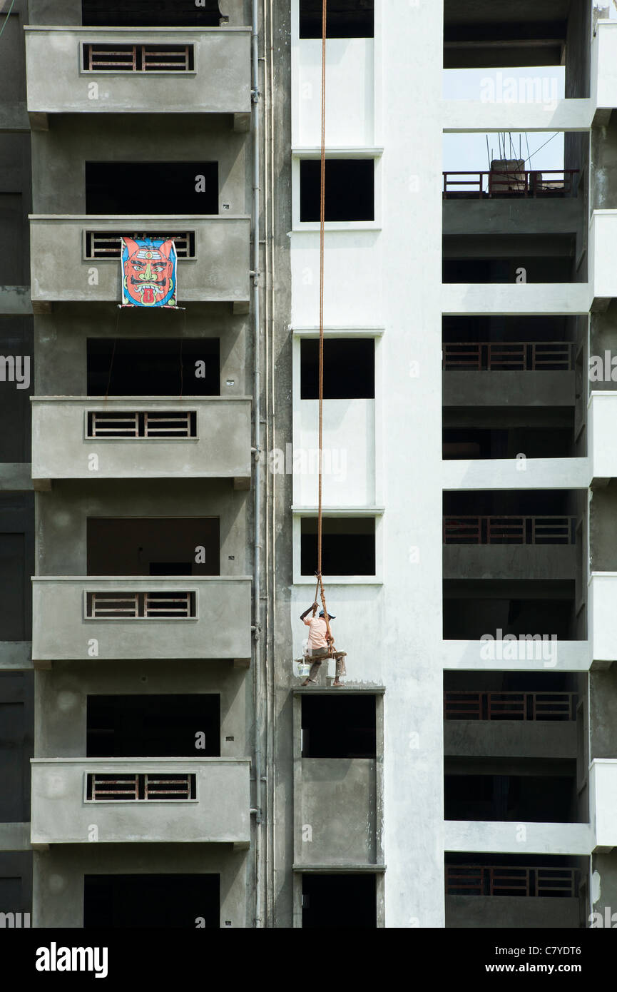Indian painter sitting on a wooden seat hanging from ropes painting the side of an apartment complex. Andhra Pradesh, India Stock Photo