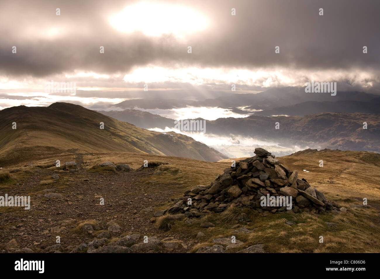 A lone cairn on the summit of the Fairfield Horseshoe Ridge and sunlight streams through the mist covered valley Stock Photo