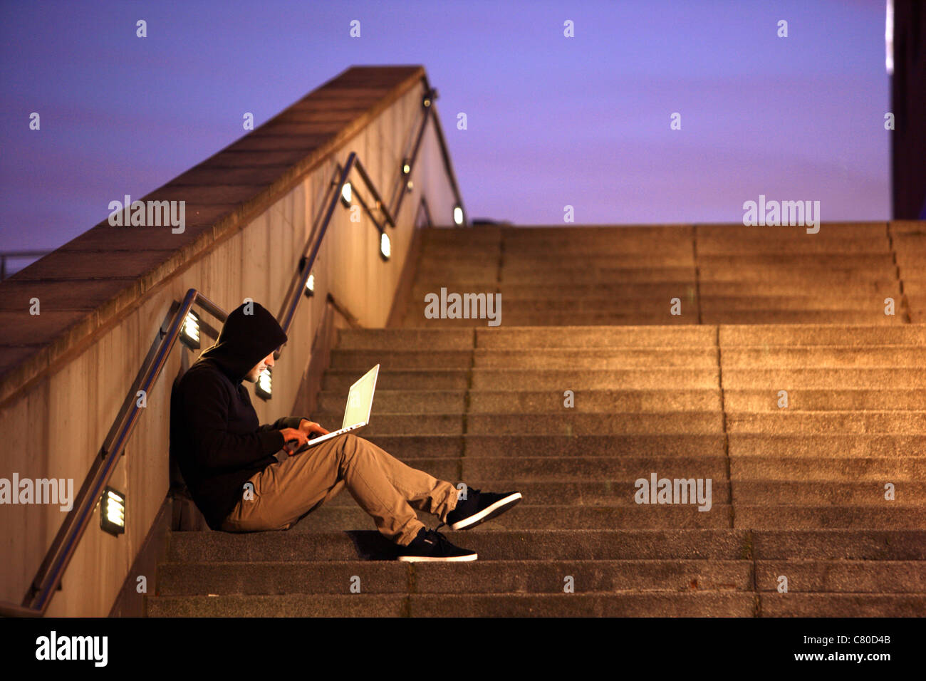 Computer user, hacker, sits conspiratorially, outside, on stairs, with a laptop. Symbol picture, computer-Internet crime. Stock Photo