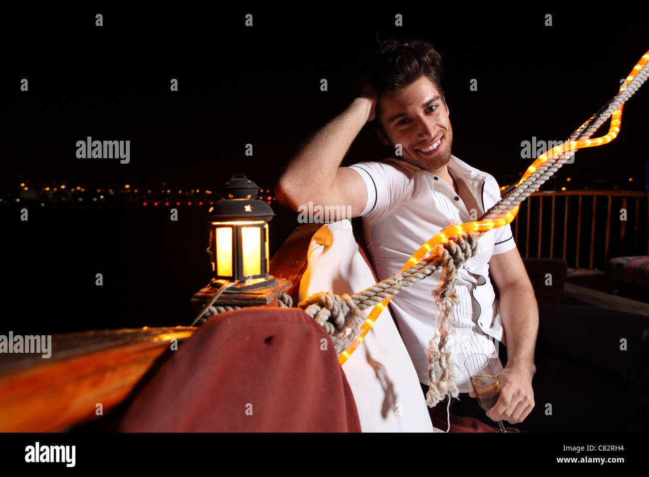 young man sitting on the boat holding champagne glass,  night lights of large city in background, laughing, feeling happy Stock Photo
