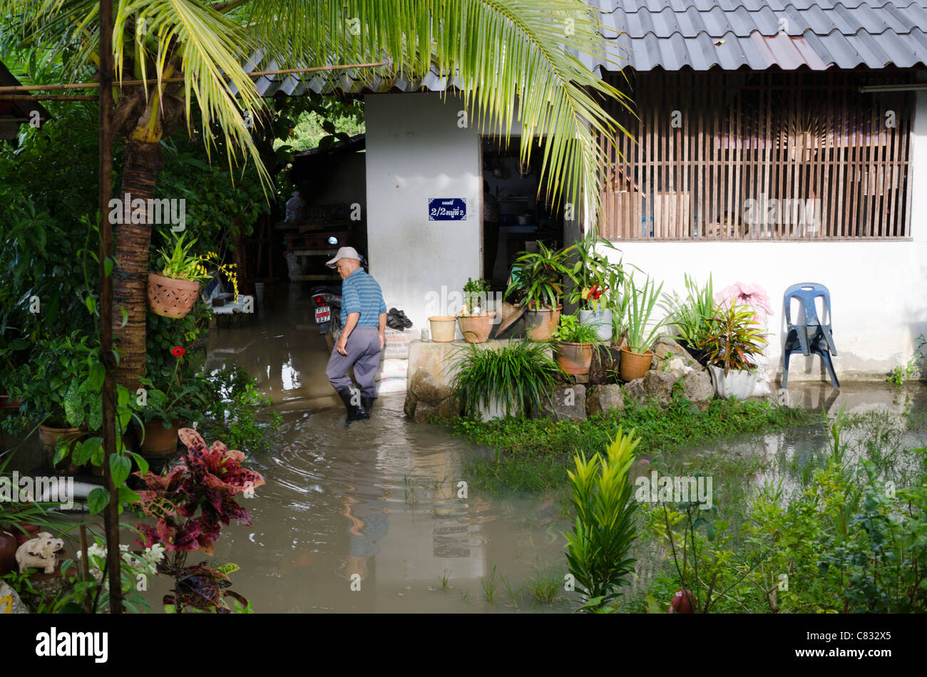 Old man wearing a hat & galoshes walks in front of house and flooded yard in Thailand after a massive thunderstorm Stock Photo