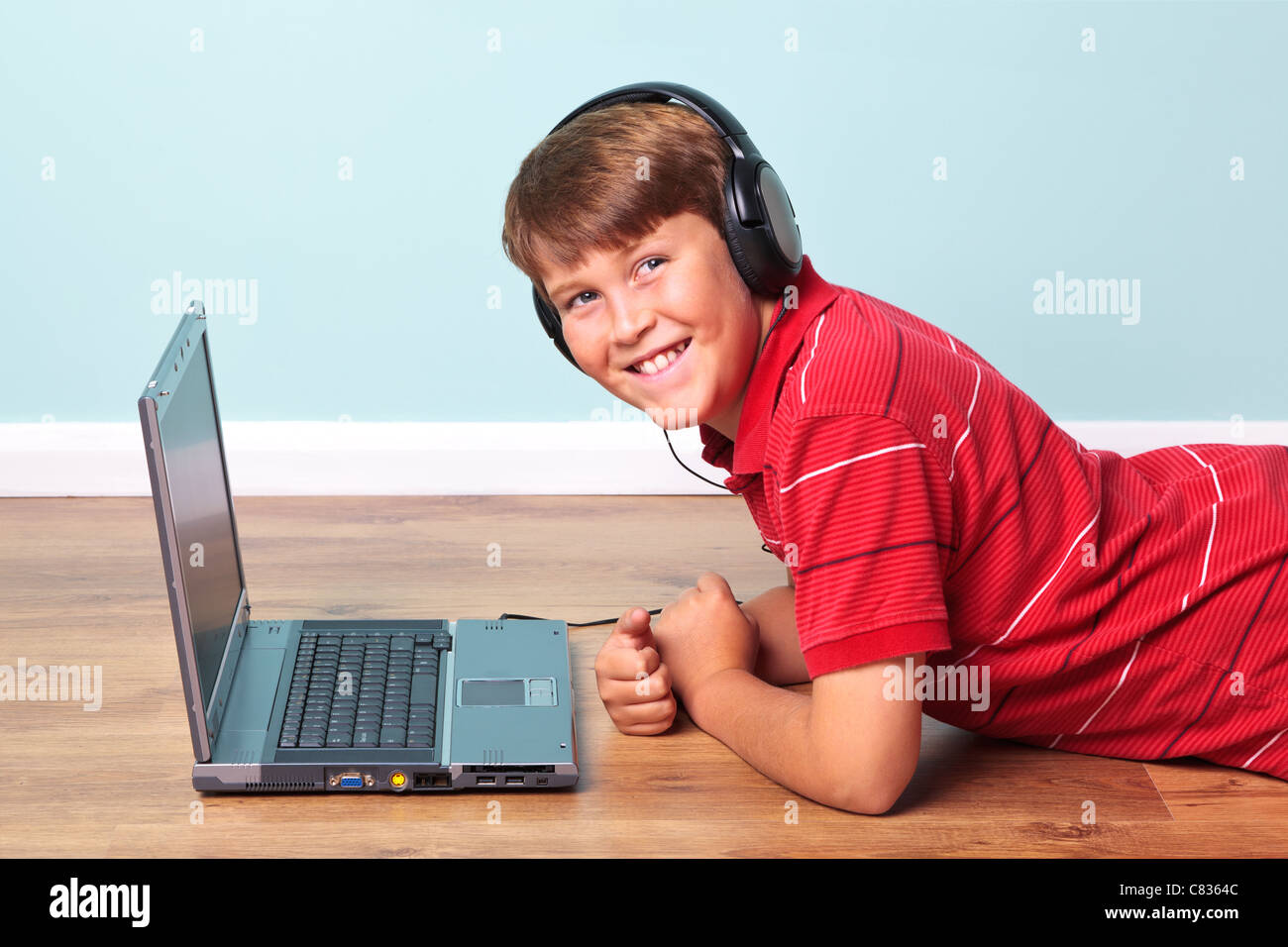 Photo of a boy wearing headphones looking towards camera lying on the floor next to his laptop computer. Stock Photo