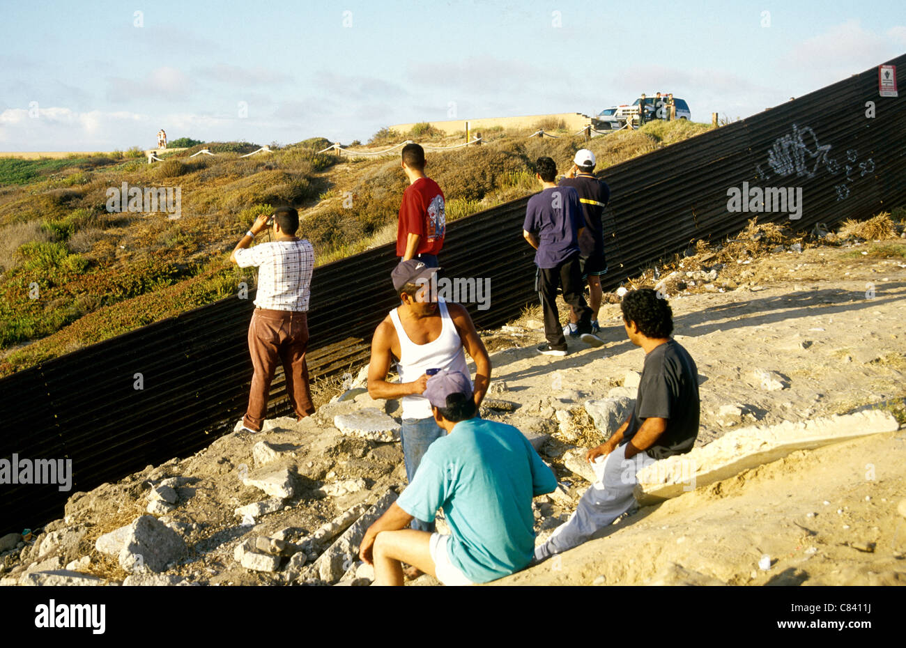 Mexican men looking at the USA  from a Tiguana beach under the watchful eyes of US border guards. Stock Photo