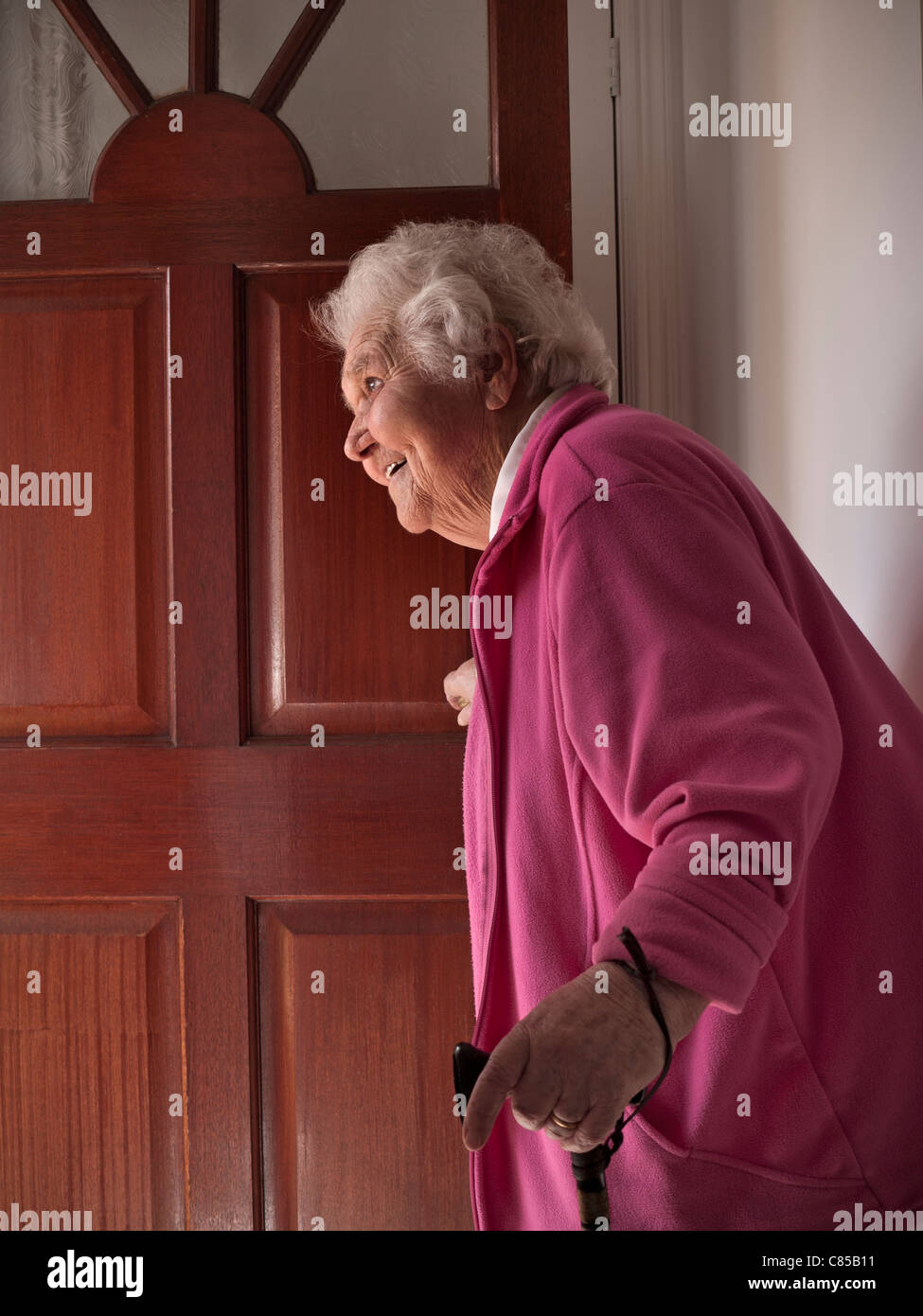 Elderly front door senior happy smiling lady standing with walking stick aid at her open front door smiling to welcome family visitor carer Stock Photo