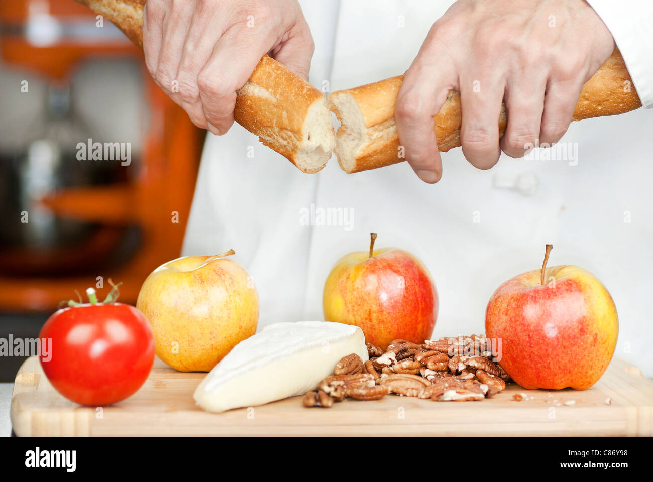 Close-up of a chef breaking a baguette over a cutting board of apples, pecans, a tomato and brie. Stock Photo