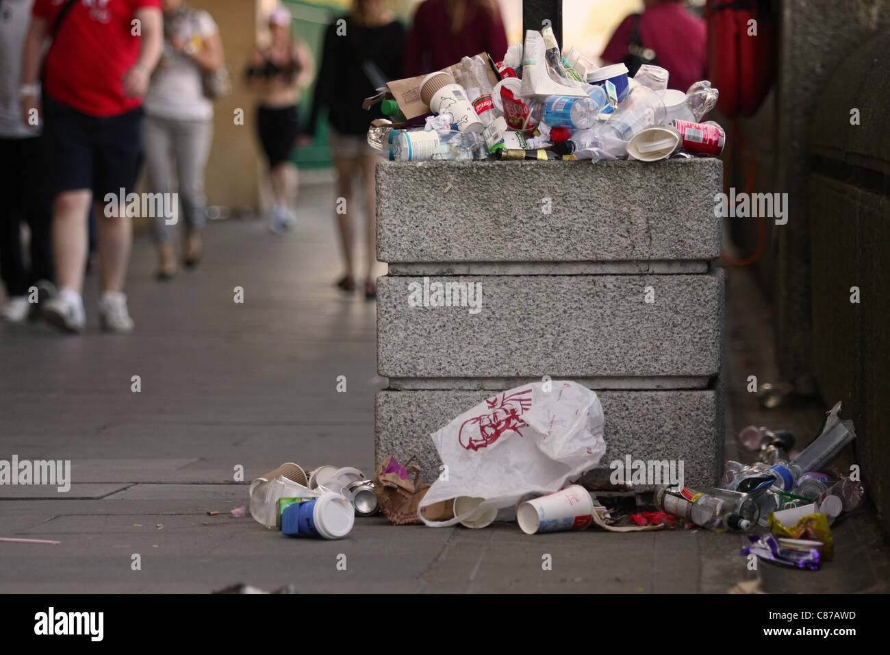 litter overflowing out of a litter bin with out of focus peole walking