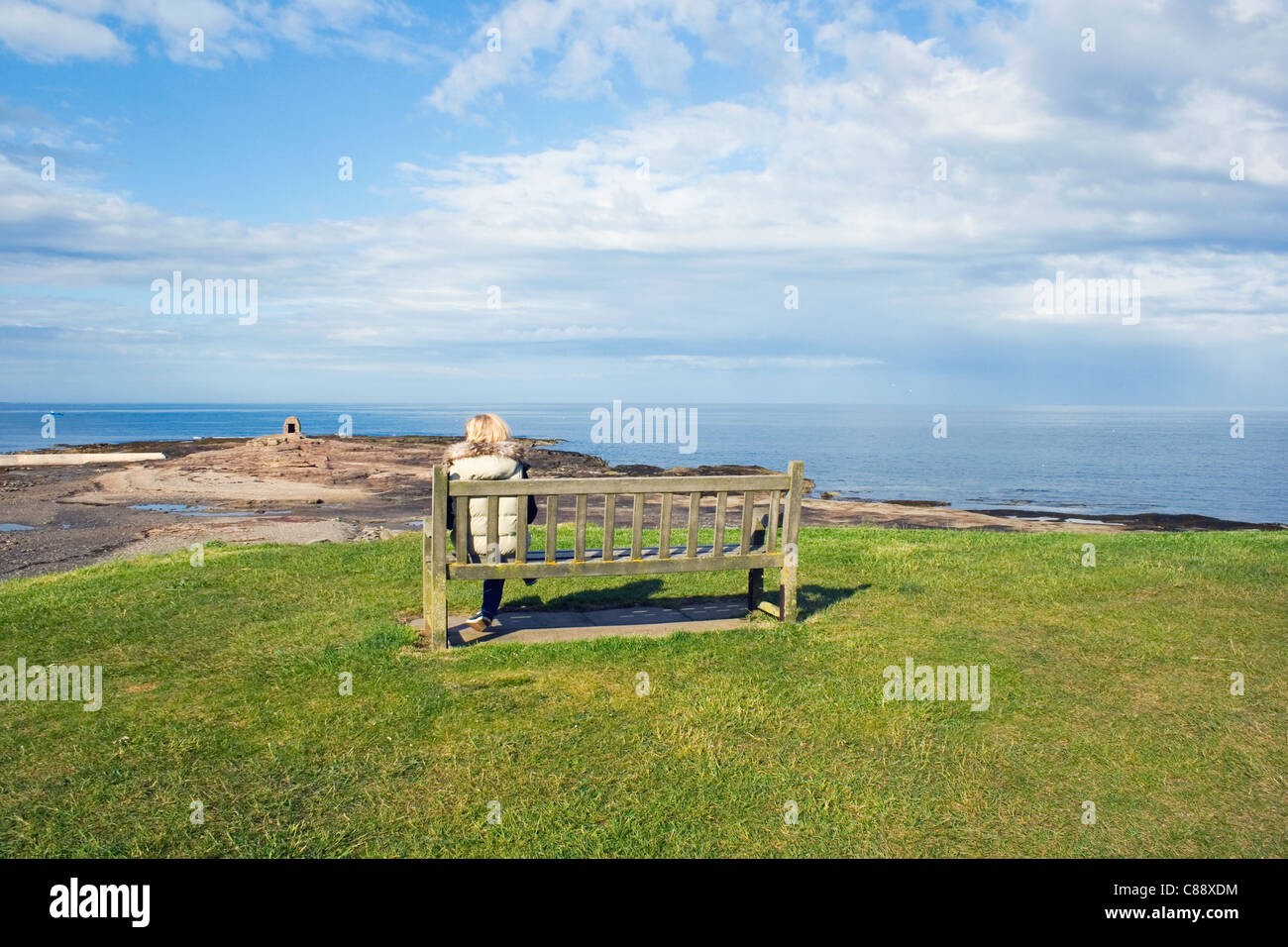 Solitary woman sitting on a bench overlooking the beach. Seahouses, Northumberland Coast, England. Stock Photo
